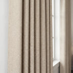 A close-up shot focusing on a light yellow-tan curtain panel hanging in crisp, neat vertical pleats. The fabric's subtle textured pattern is highlighted against a bright white window frame in the background.
