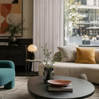 A stylish modern lounge interior featuring a black round coffee table in the foreground, a teal accent chair on the left, and an off-white curtain panel softly framing a large window behind a cream sofa