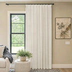 A bright, cozy living room scene featuring a single panel of off-white woven curtains hanging from a black rod next to a window. A white textured ottoman holding a potted green plant sits in front of the window, with a partial view of a beige sofa and a framed abstract painting on the wall.