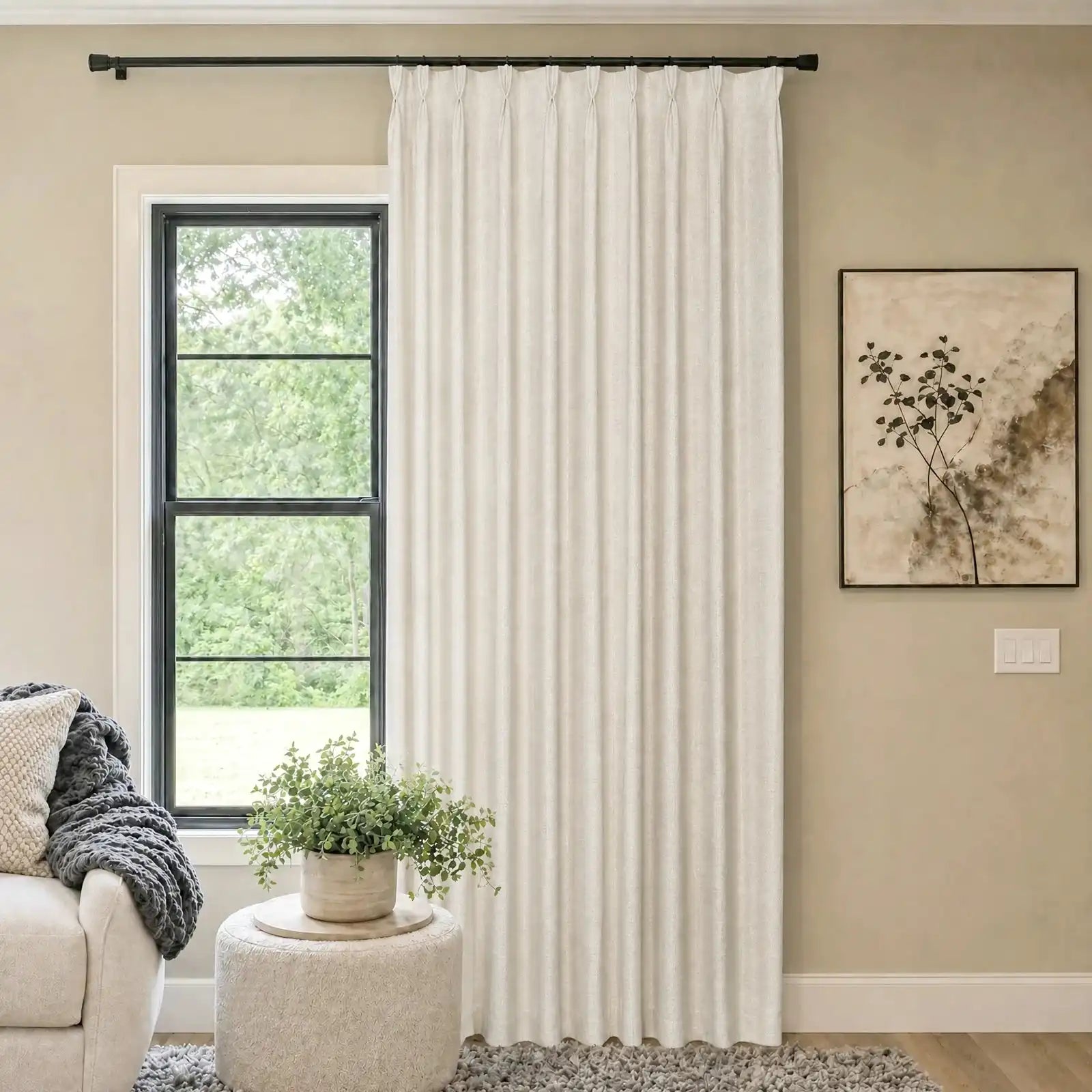 A bright, cozy living room scene featuring a single panel of off-white woven curtains hanging from a black rod next to a window. A white textured ottoman holding a potted green plant sits in front of the window, with a partial view of a beige sofa and a framed abstract painting on the wall.
