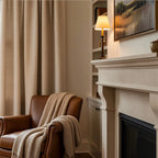 A cozy living room corner featuring a classic cream-colored fireplace mantel on the right with a small illuminated lamp. To the left, a textured tan-yellow curtain hangs beside a brown leather armchair draped with a thick, chunky knit beige blanket.
