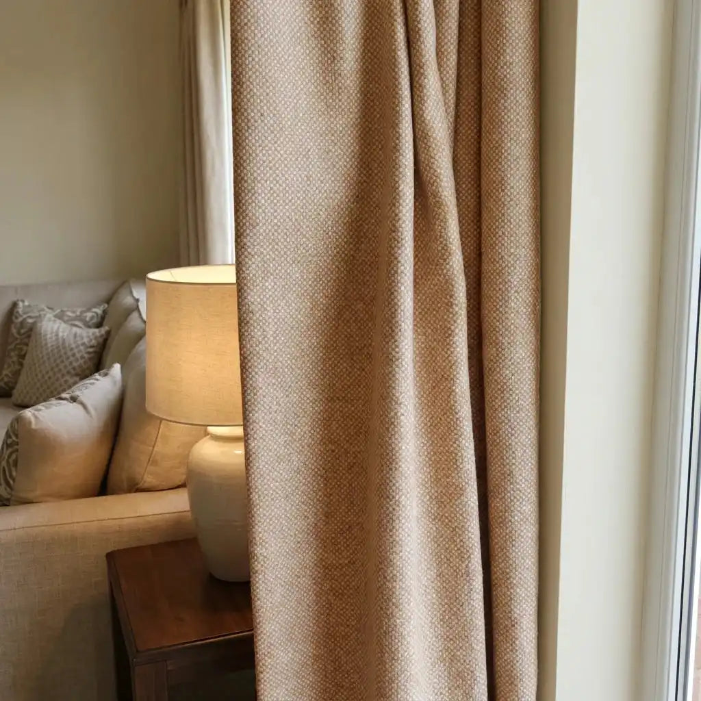 A close-up interior shot of a mustard-yellow textured curtain panel softly draped next to a white ceramic table lamp on a dark wood end table. Part of a cream-colored sofa with patterned pillows is visible in the background.