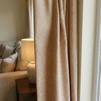 A close-up interior shot of a mustard-yellow textured curtain panel softly draped next to a white ceramic table lamp on a dark wood end table. Part of a cream-colored sofa with patterned pillows is visible in the background.